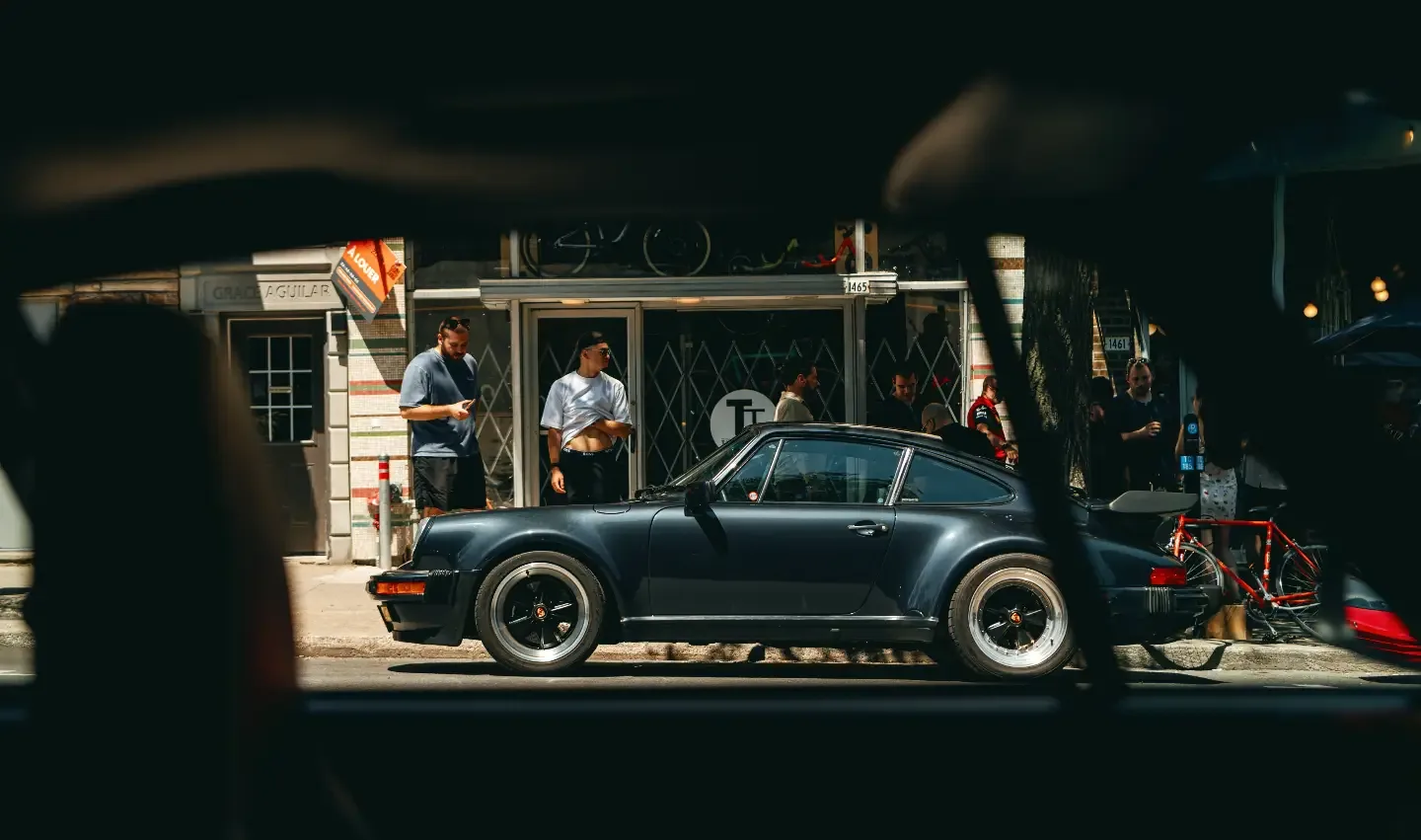 A black Porsche 911 Turbo in the shade of a storefront, bicycles and conversation behind it.