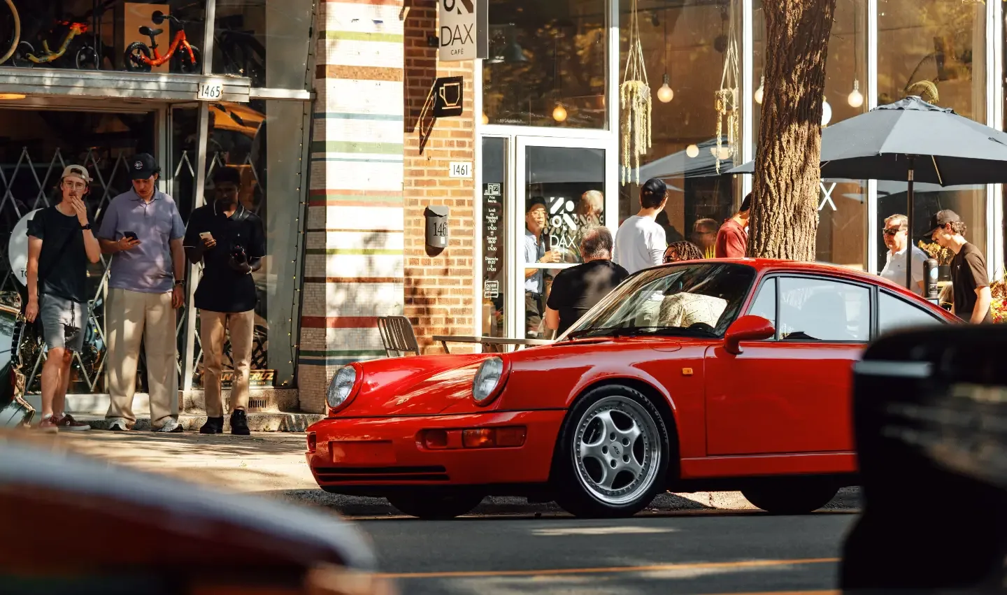 A red air-cooled Porsche 911 parked outside DAX Café while people gather on the terrace.