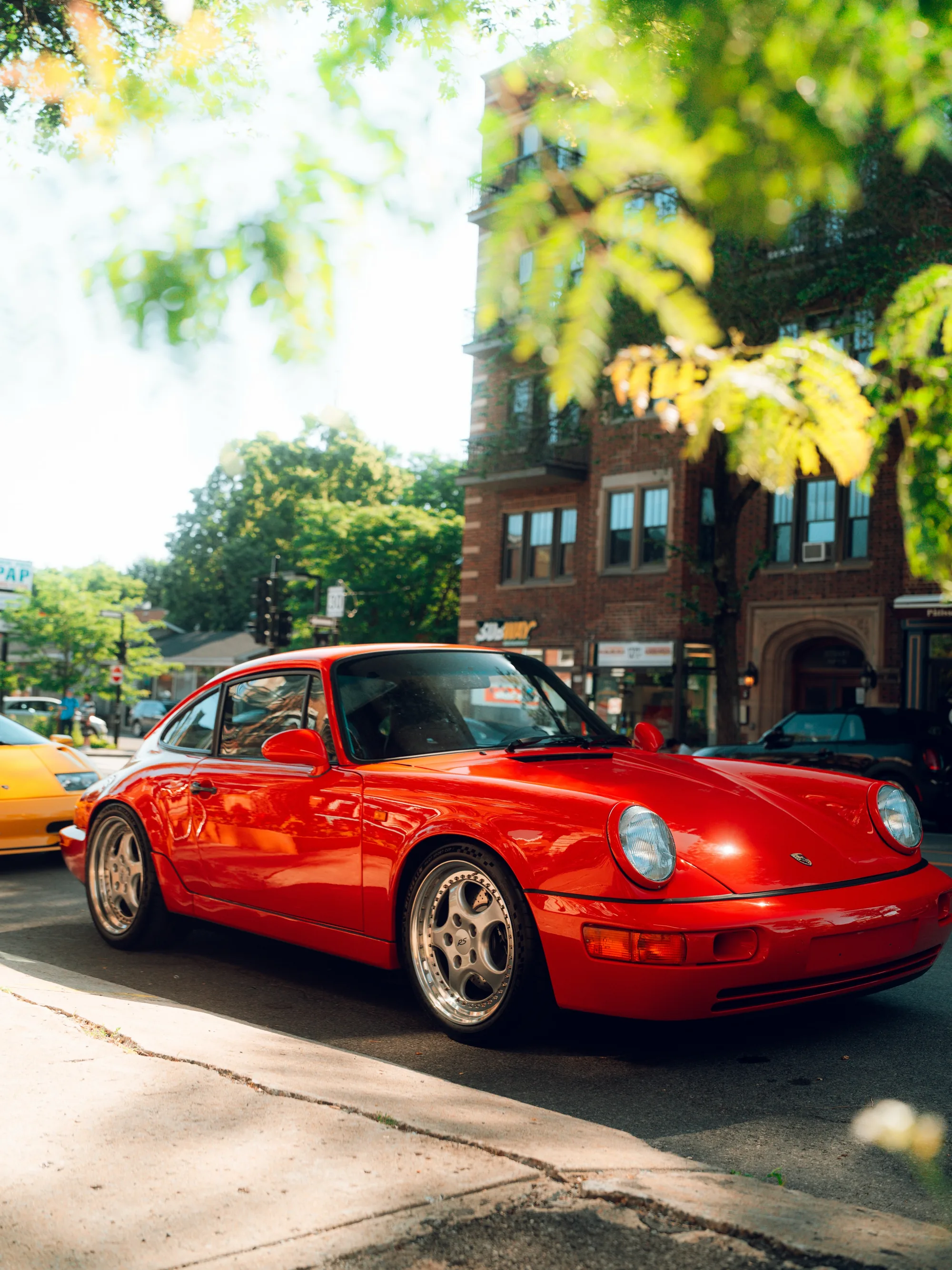 A red air-cooled Porsche 911 parked under the trees on a residential stretch.