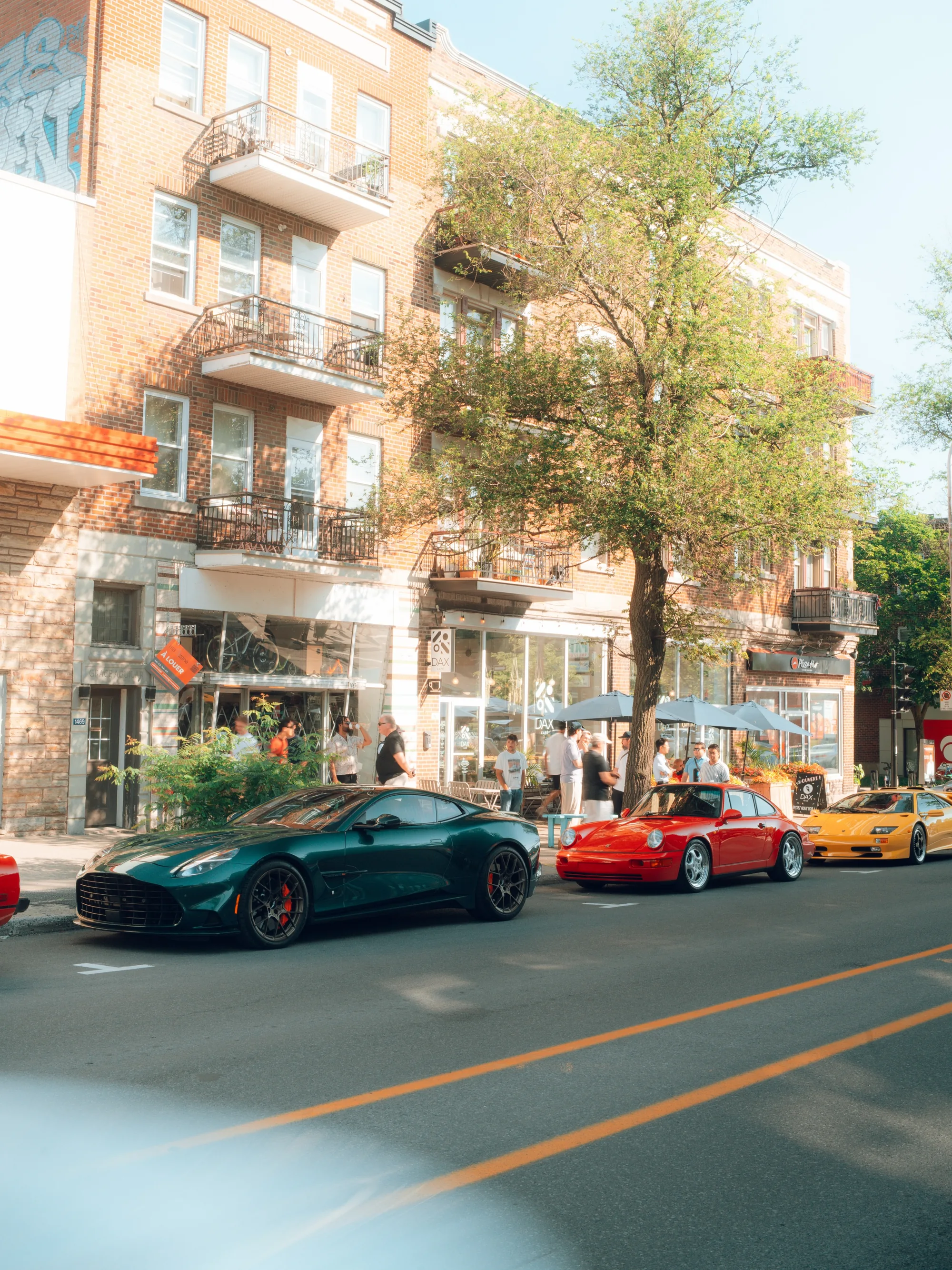 A green Aston Martin, a red sports car and a yellow Lamborghini Diablo parked along Avenue Van Horne in front of Montréal brick triplexes.