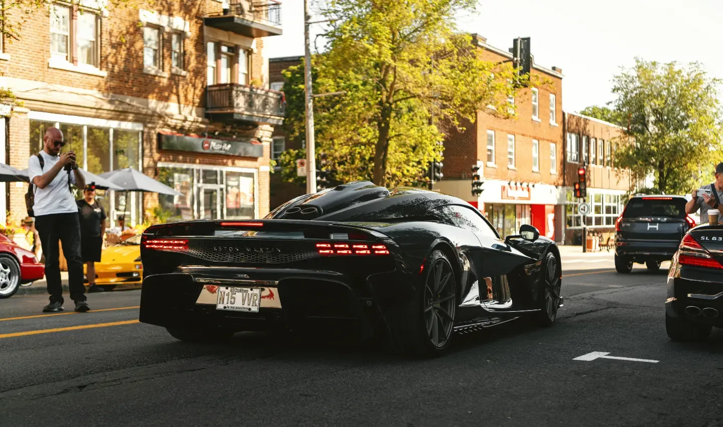 A black hypercar pulls away down the avenue as onlookers watch from the sidewalk.