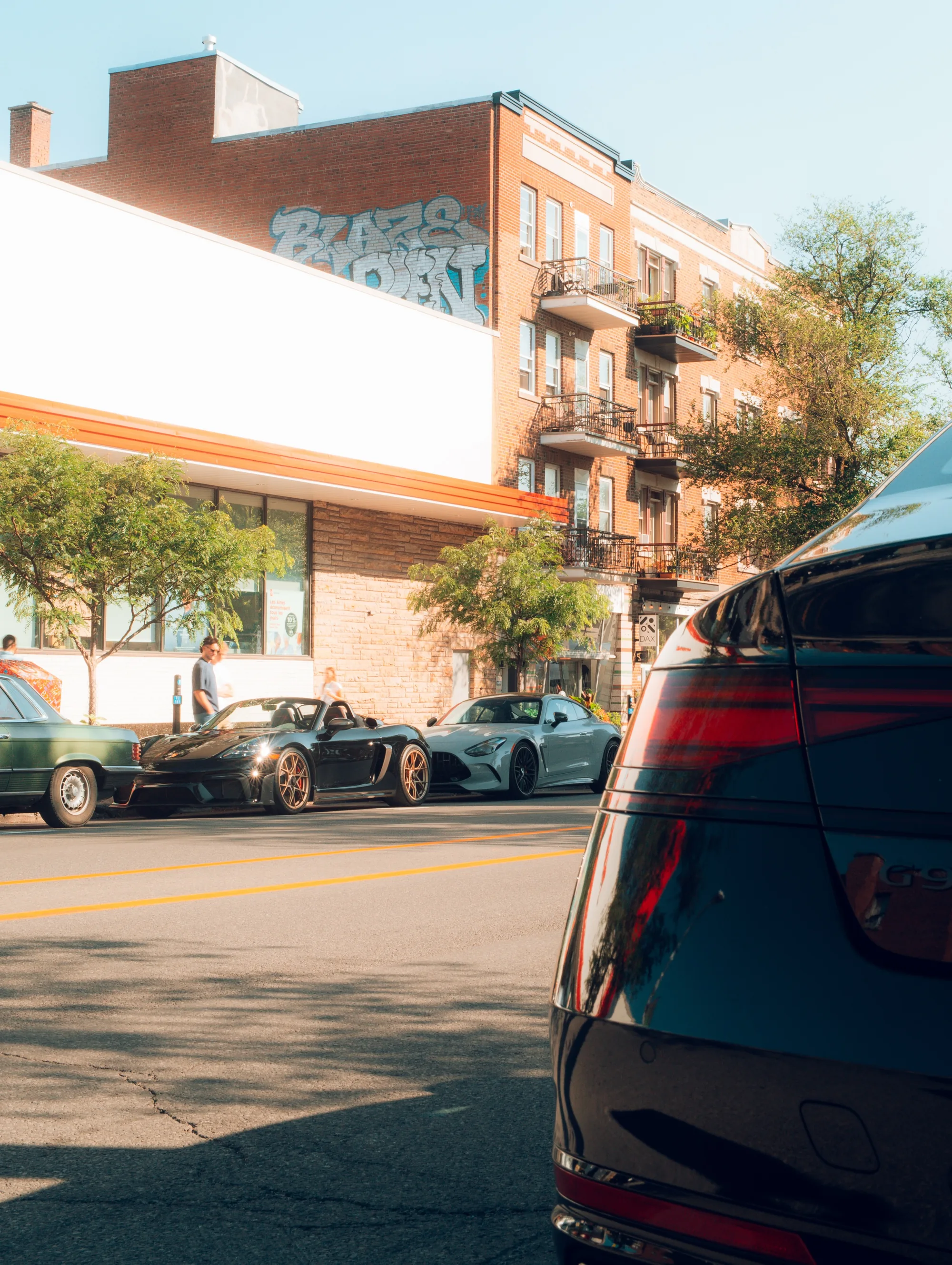 The avenue at mid-morning: an orange-awninged corner, brick triplexes, and the tail of a dark sports car in the foreground.
