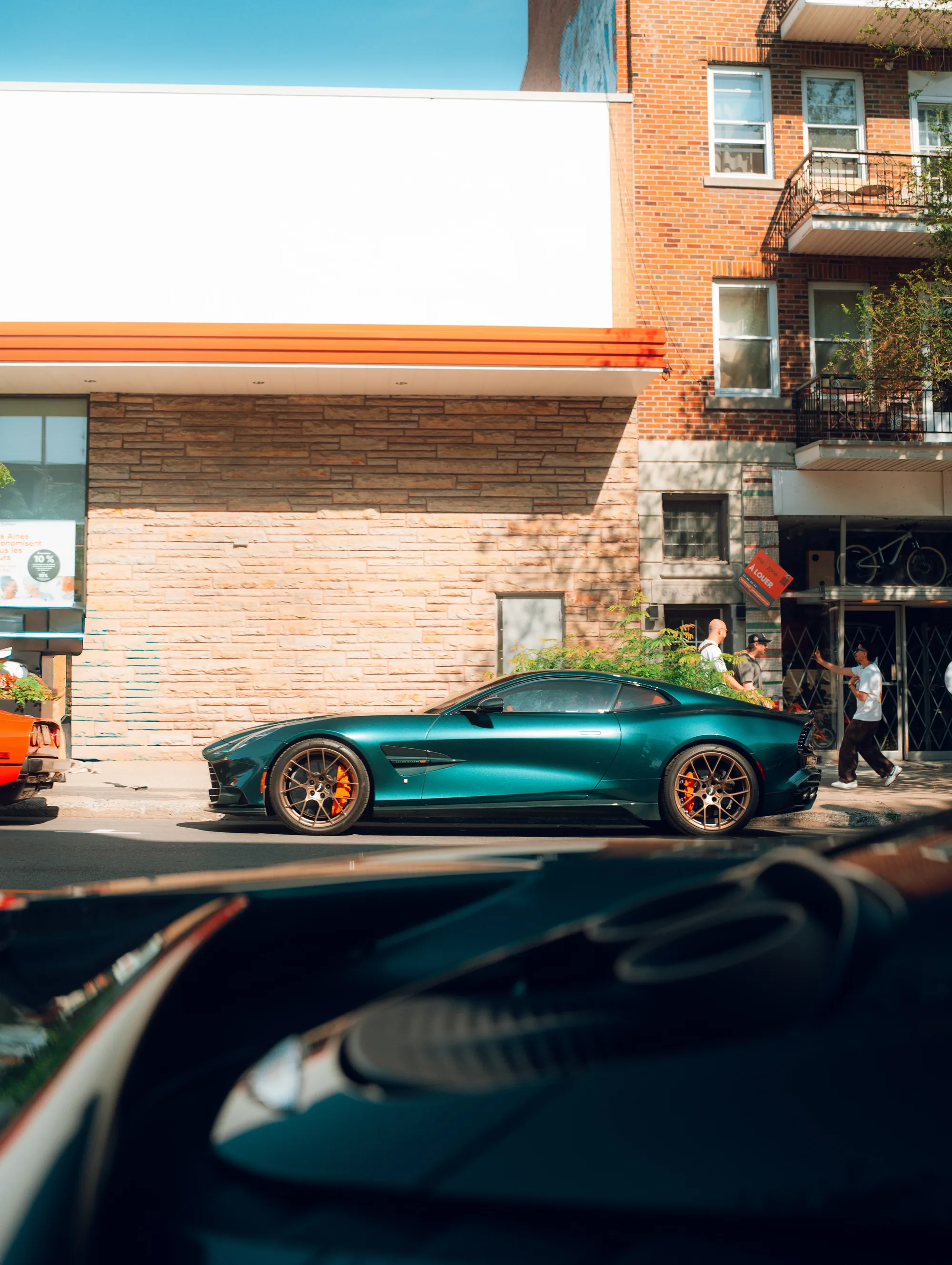 A teal Aston Martin in profile, seen over the hood of a parked car across the street.