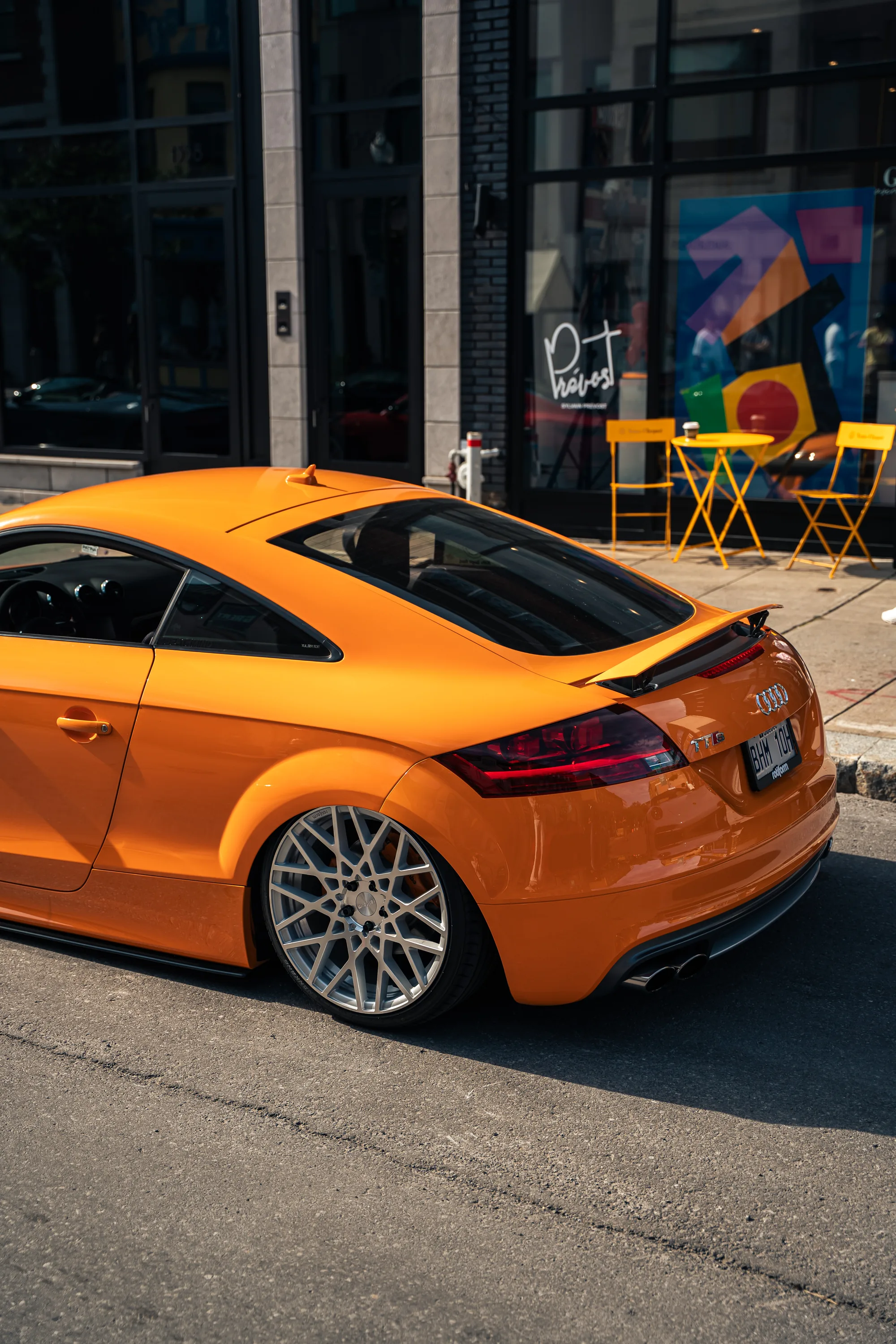 An orange Audi TT at the curb beside a café’s yellow sidewalk chairs.