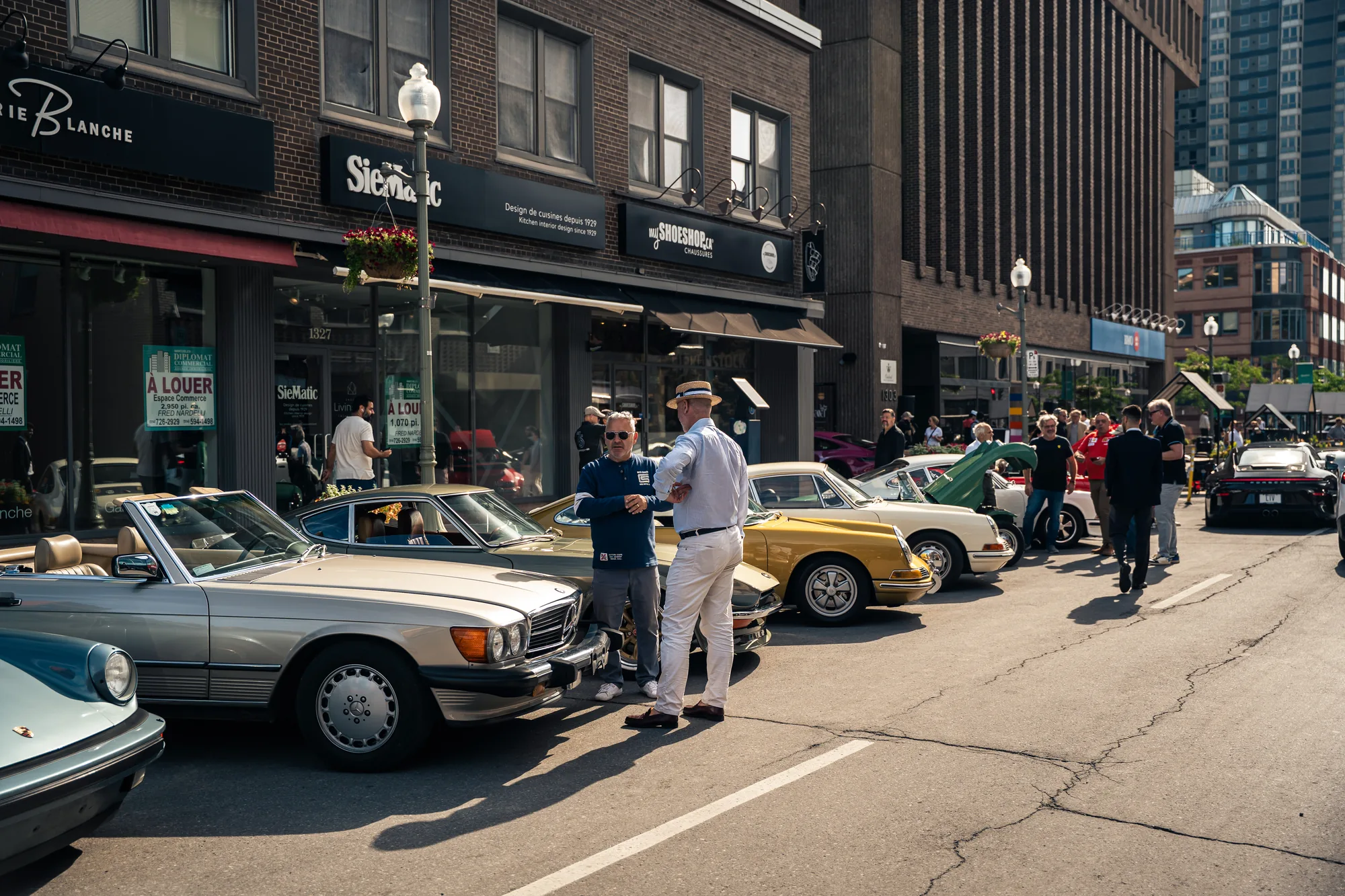 The crowd walks the middle of Greene Avenue between a silver Mercedes-Benz SL and a yellow Porsche 911.